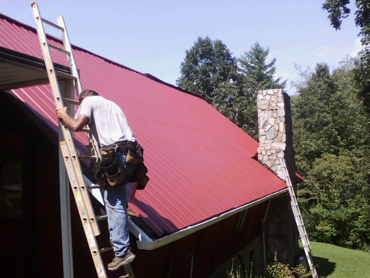 Roofer on a ladder beside a red metal roof and stone chimney — Soffit & Fascia Repair by Roofing Brothers in Brush Creek